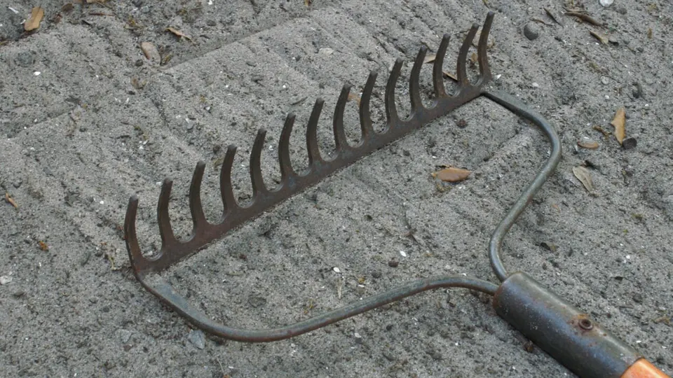 Ancinho de metal apoiado em chão de cascalho, visto de cima em luz de dia